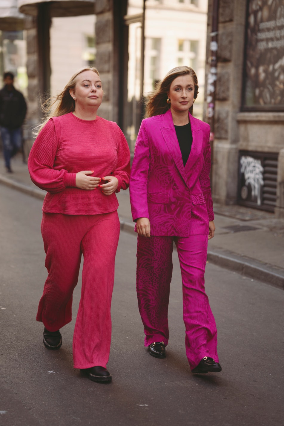 Maria Gudme og Camilla Søe går sammen på en gade. Maria Gudme har hænderne samlet foran sig. Hun er i et rødligt sæt, Camilla Søe har et pink jakkesæt på. 